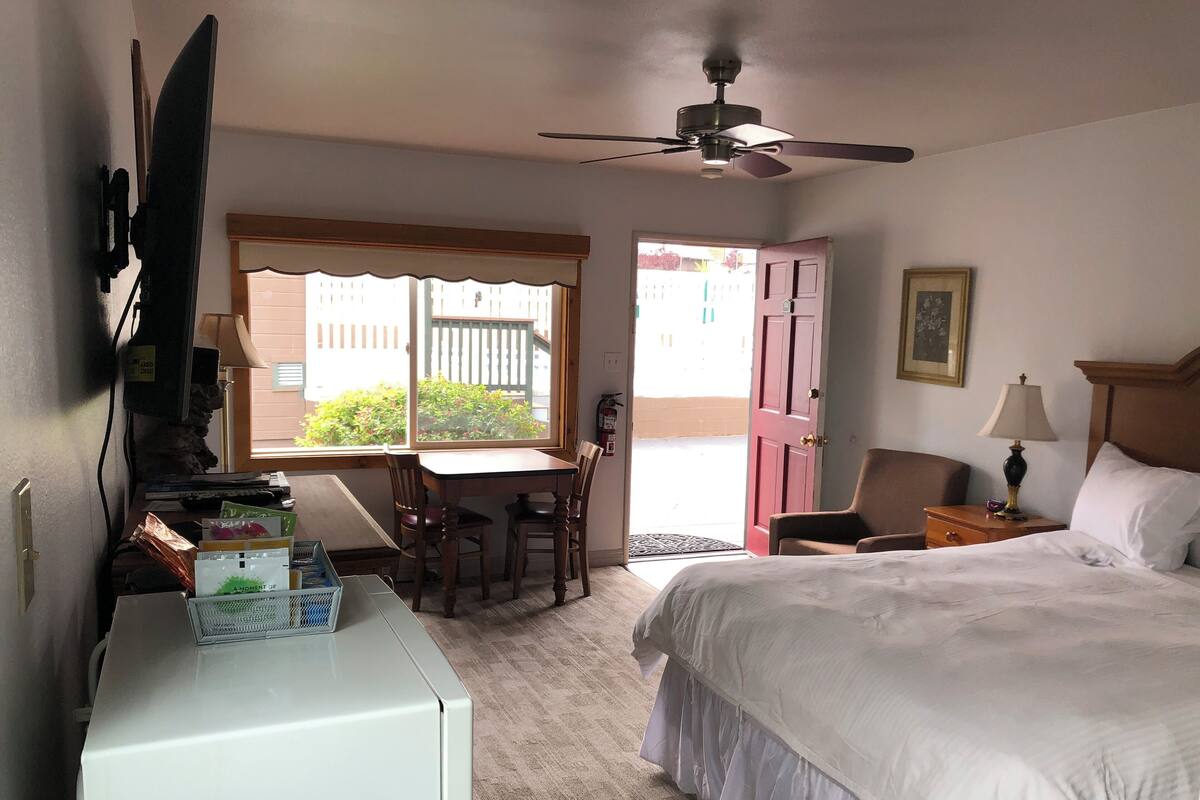 A hotel room with a bed, a desk, a chair, a ceiling fan, and a window with a view of the outside.