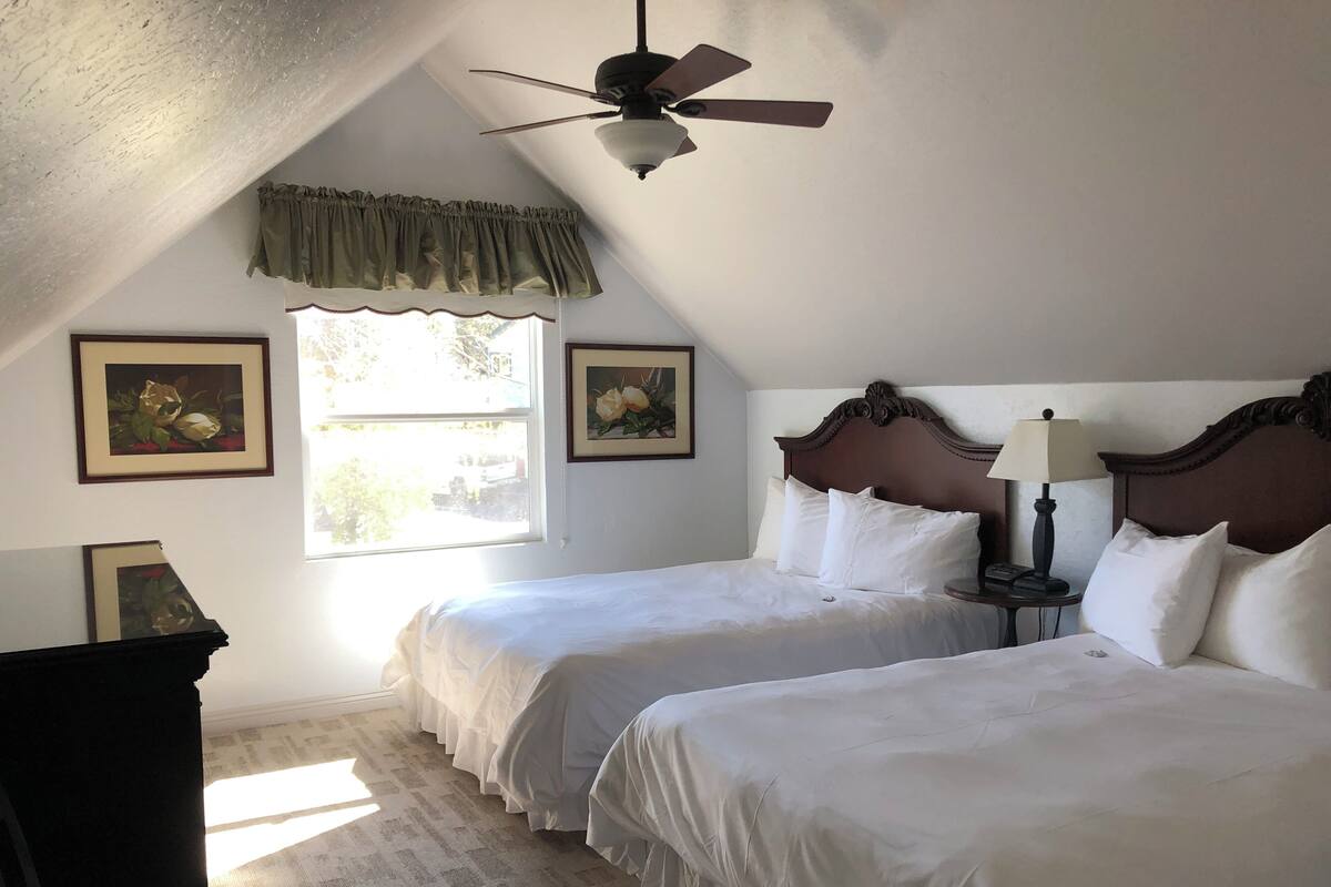 A bedroom with two beds, a ceiling fan, a window with curtains, and framed artwork on the walls.
