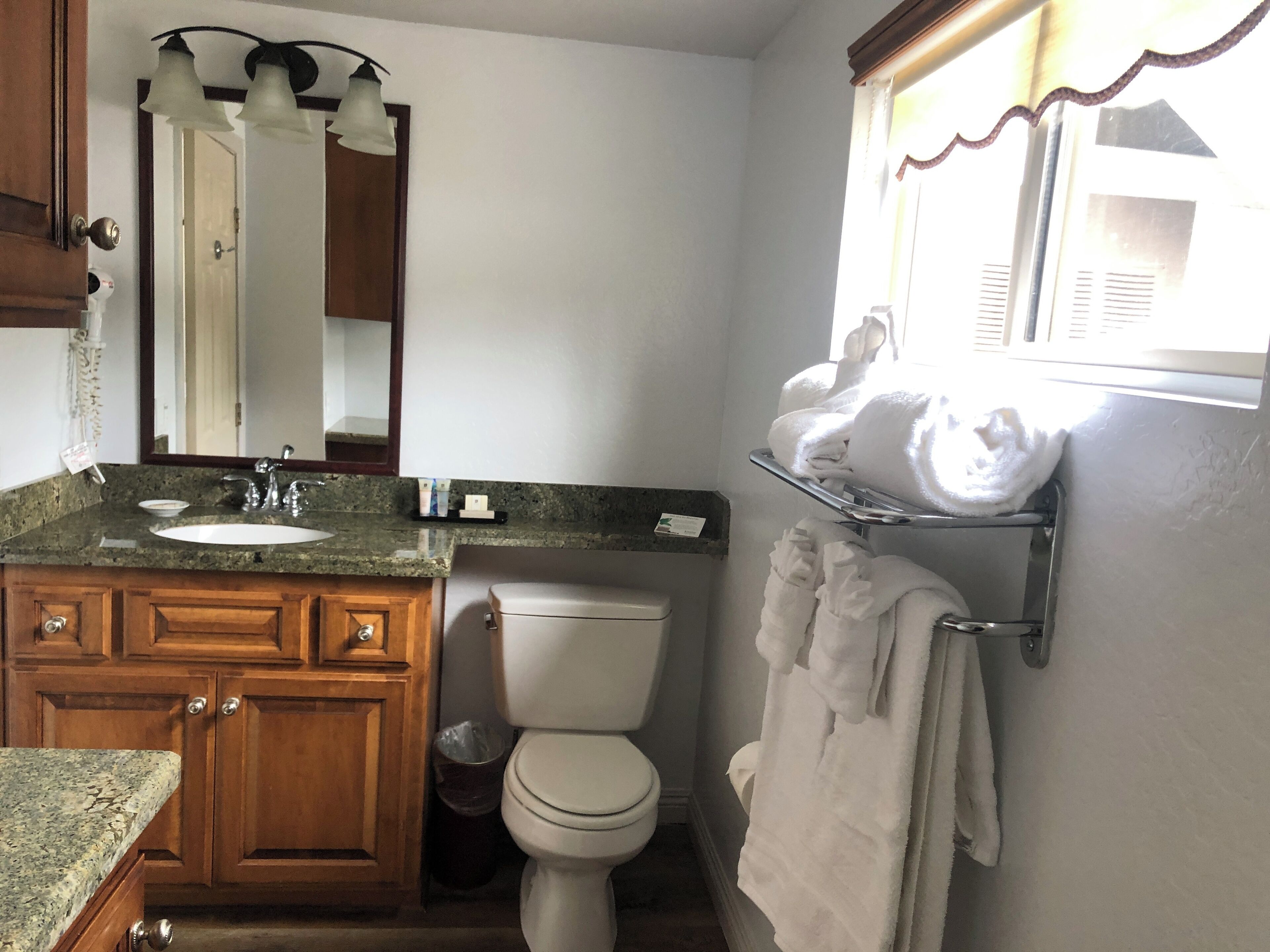 A bathroom with a granite countertop sink, wooden cabinets, a white toilet, a mirror with three lights, and a towel rack with towels.