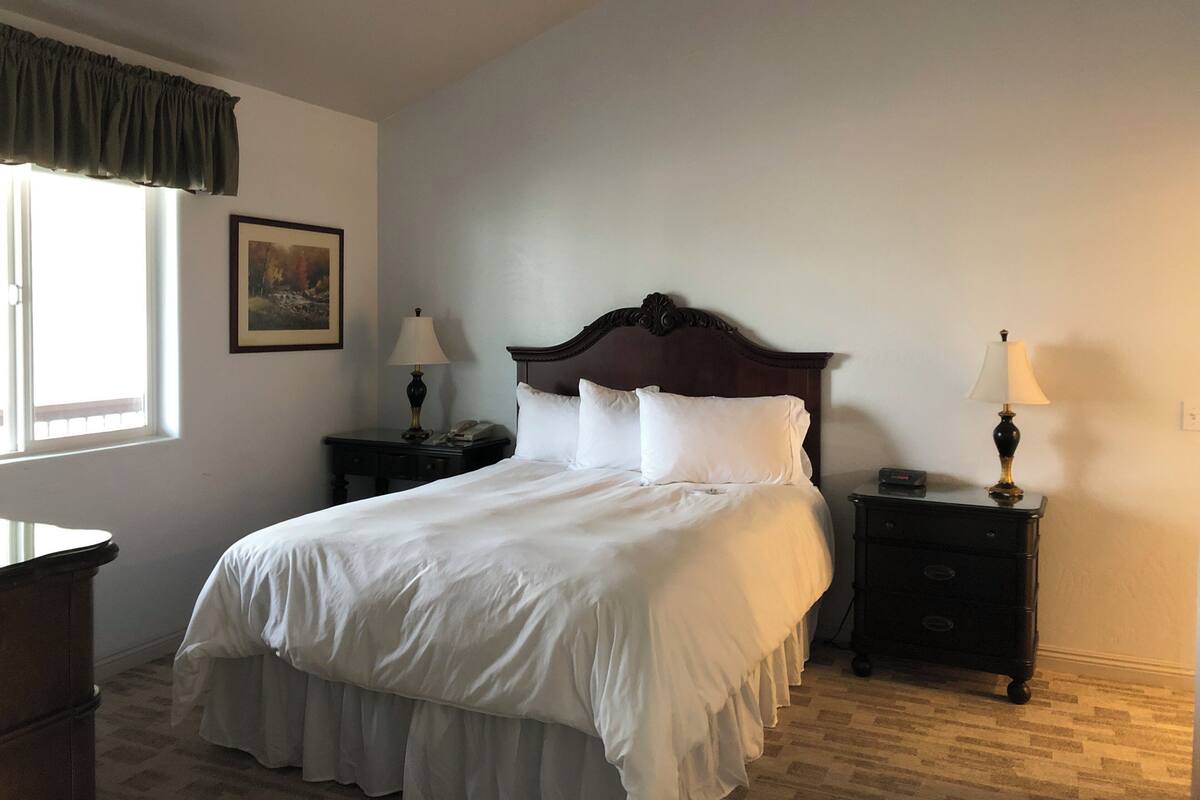 A neatly made bed with white linens, two bedside tables with lamps, a framed picture on the wall, and a window with curtains.