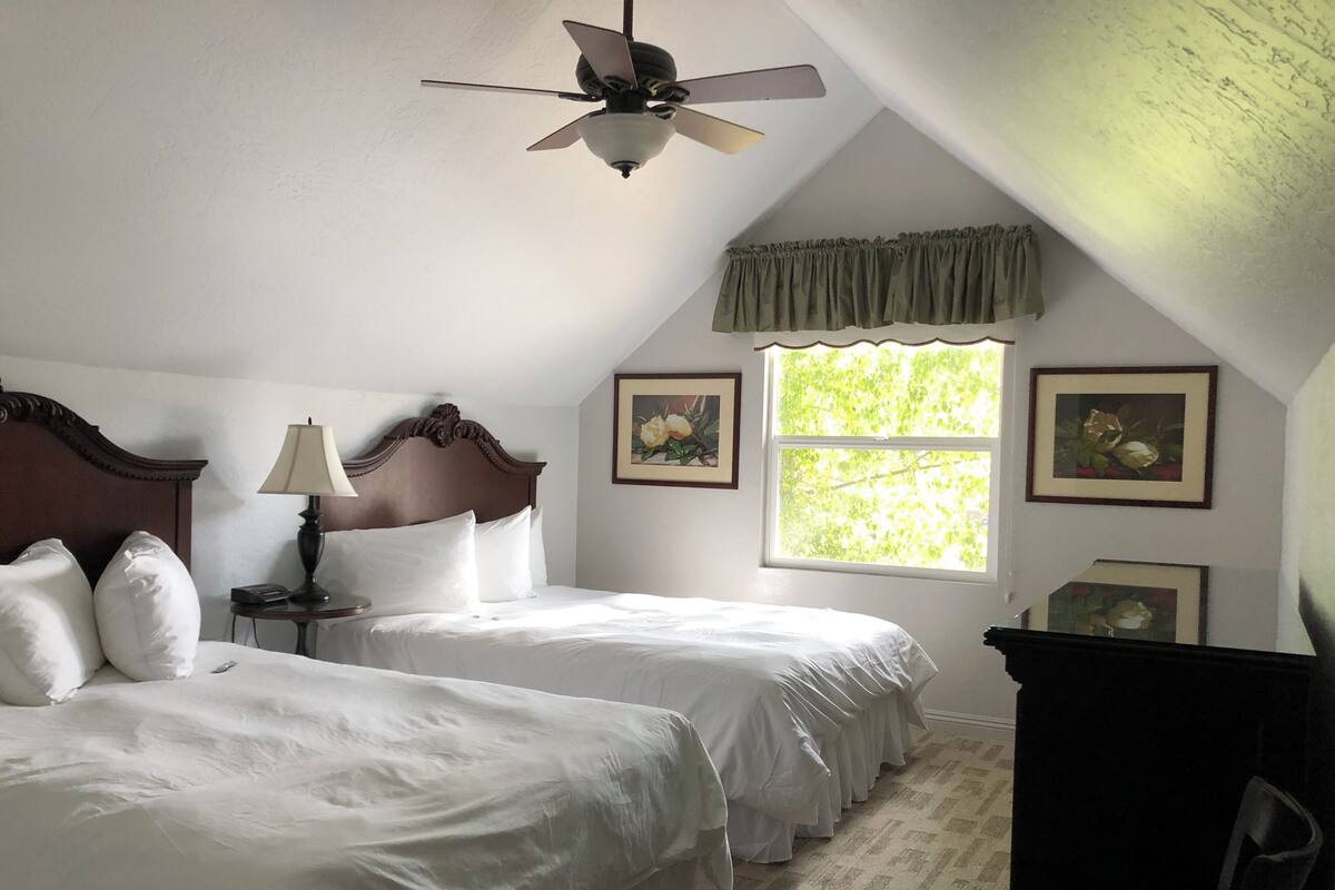 A bedroom with two beds, a ceiling fan, a window with a view of greenery, and framed pictures on the wall.