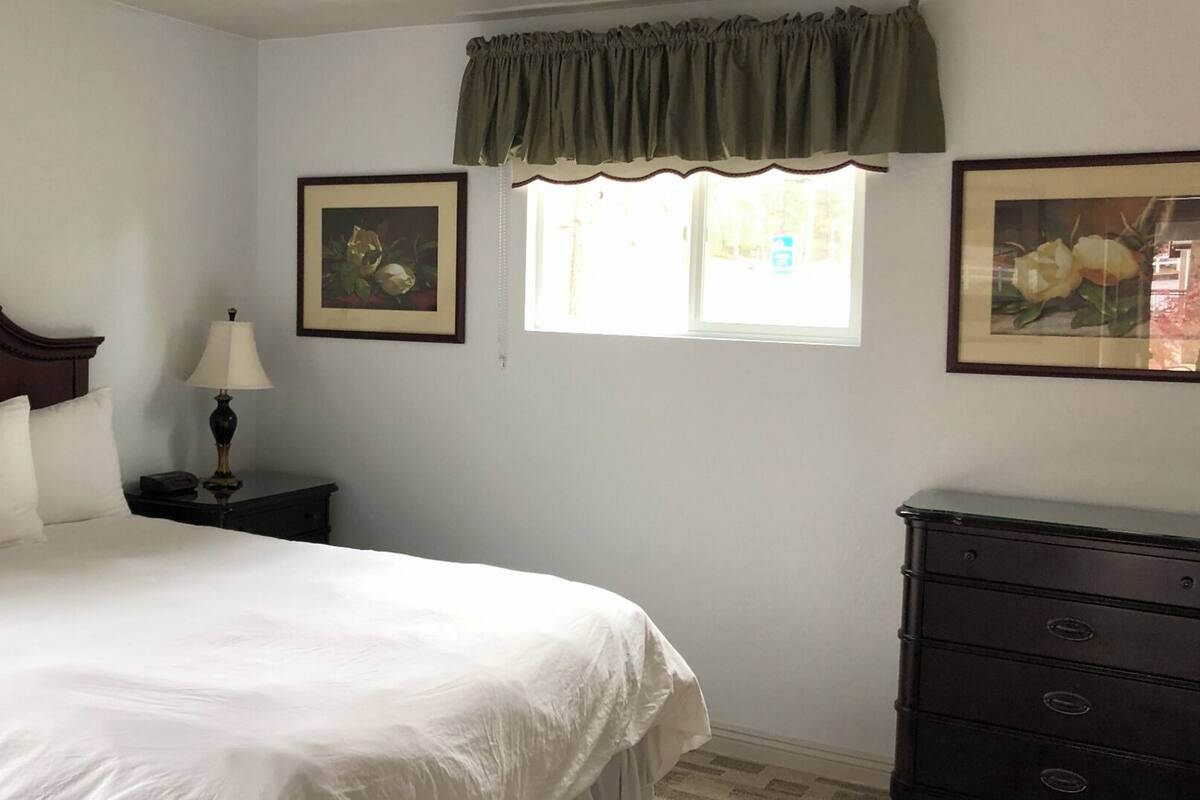 A neatly made bed with white linens, a wooden headboard, and a nightstand with a lamp. There is a window with a green valance and two framed pictures on the wall.