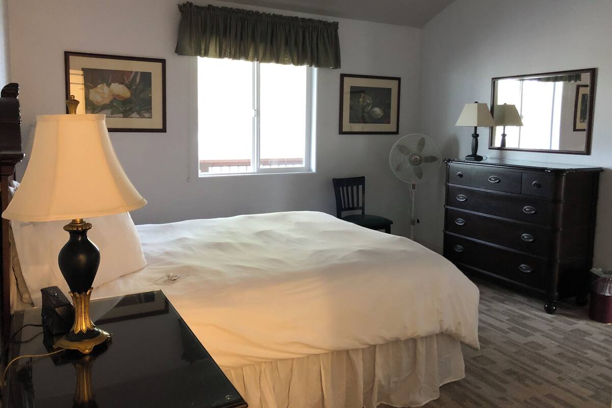 A bedroom with a bed, a dresser, a desk with a lamp, a chair, a window, and two wall-mounted fans.