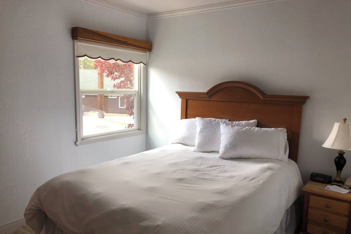 A neatly made bed with white linens in a room with a wooden headboard, a bedside table with a lamp, and a window with a view of trees.