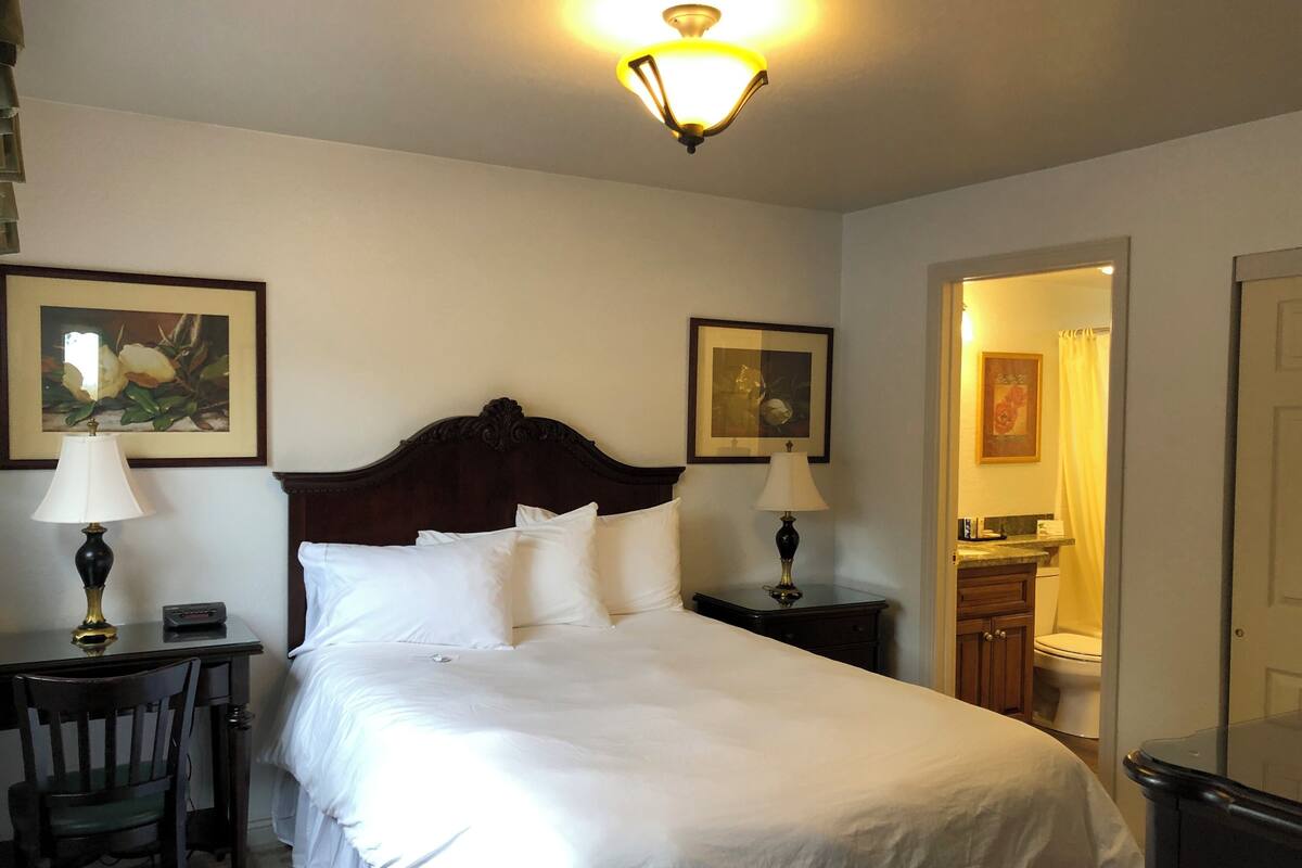 A neatly made bed with white linens, two bedside lamps, a wooden nightstand, a chair, and a painting on the wall.