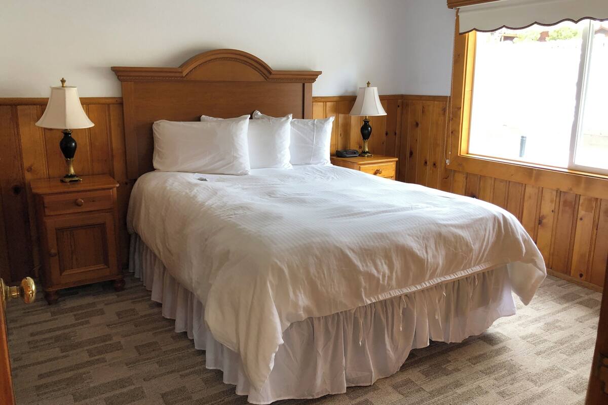 A neatly made bed with white linens, two bedside tables with lamps, and a window with a view of greenery.