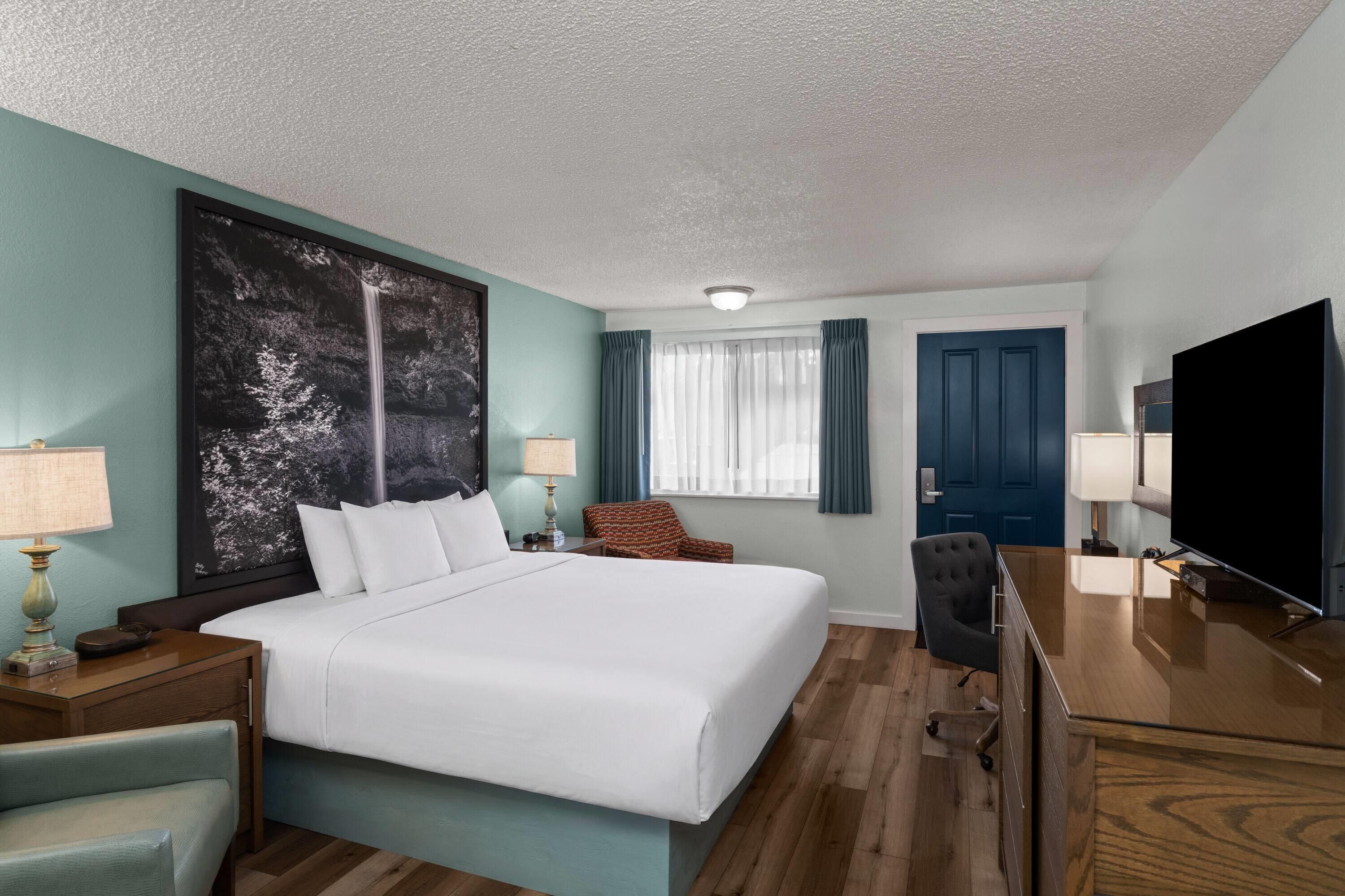 Habitación de hotel con cama, escritorio con televisor, silla, lámpara y un cuadro grande en la pared.