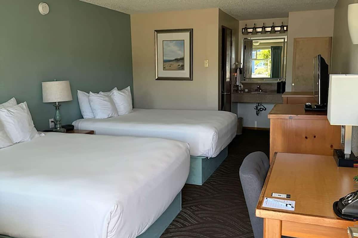 A hotel room with two beds, a desk, a chair, a lamp, a mirror, and a window with a view of the outdoors.