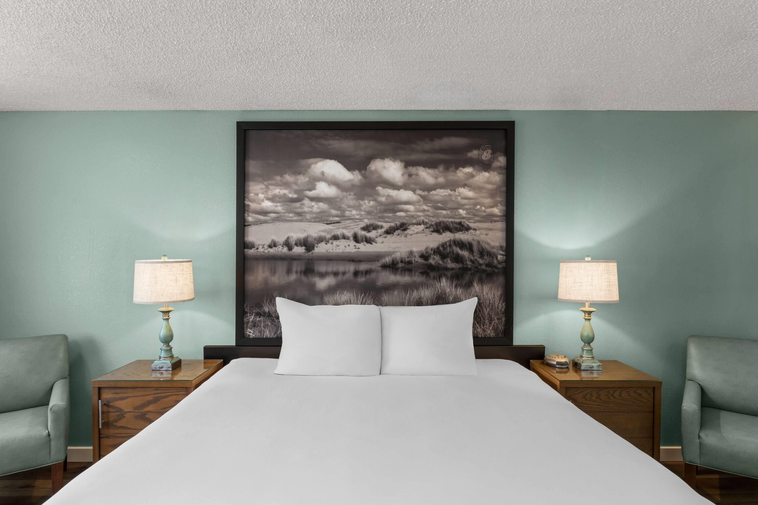 A hotel room with a large bed, two nightstands, two lamps, and a wall-mounted picture of a landscape.