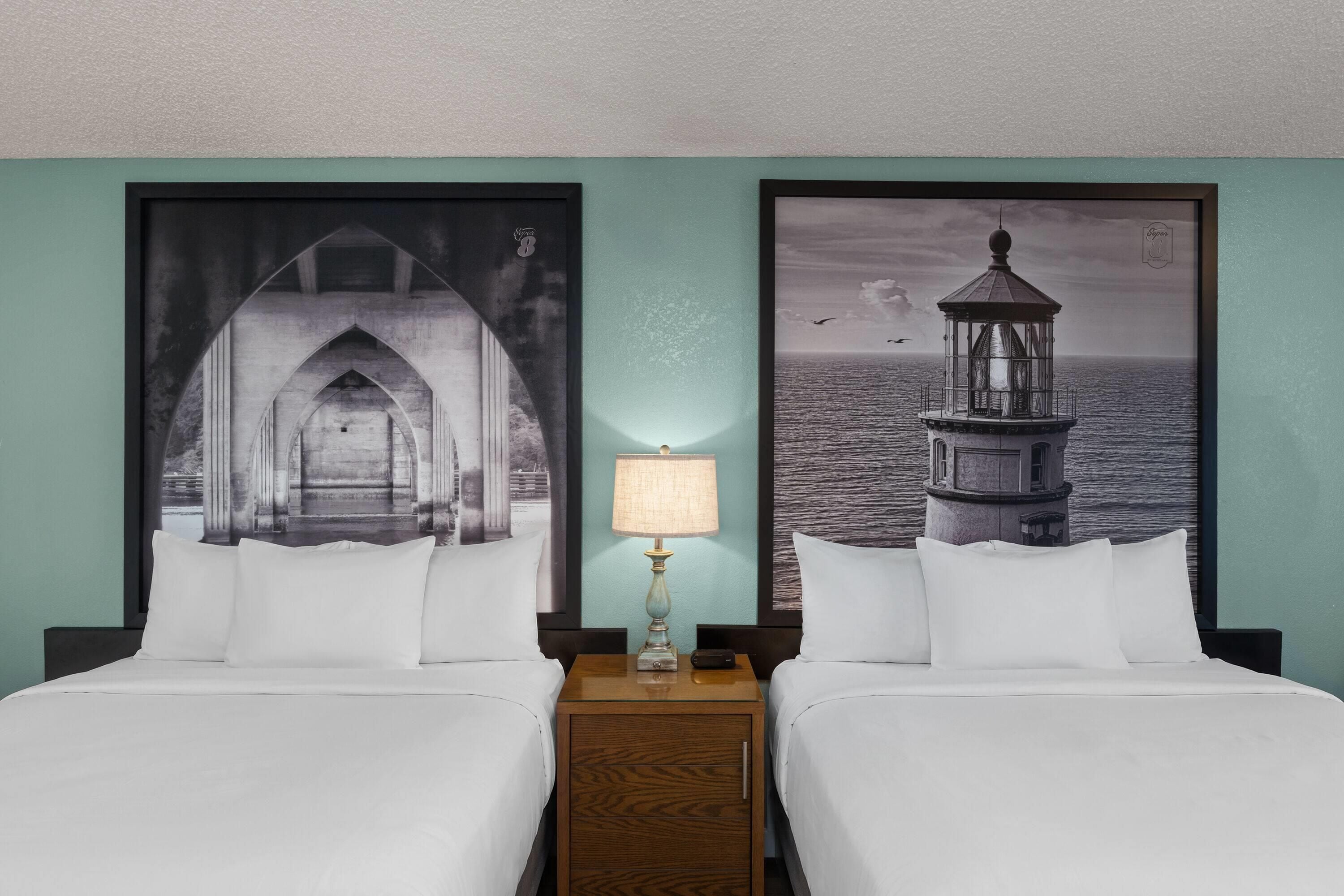 A hotel room with two beds, a nightstand, a lamp, and two large framed pictures of a bridge and a lighthouse.