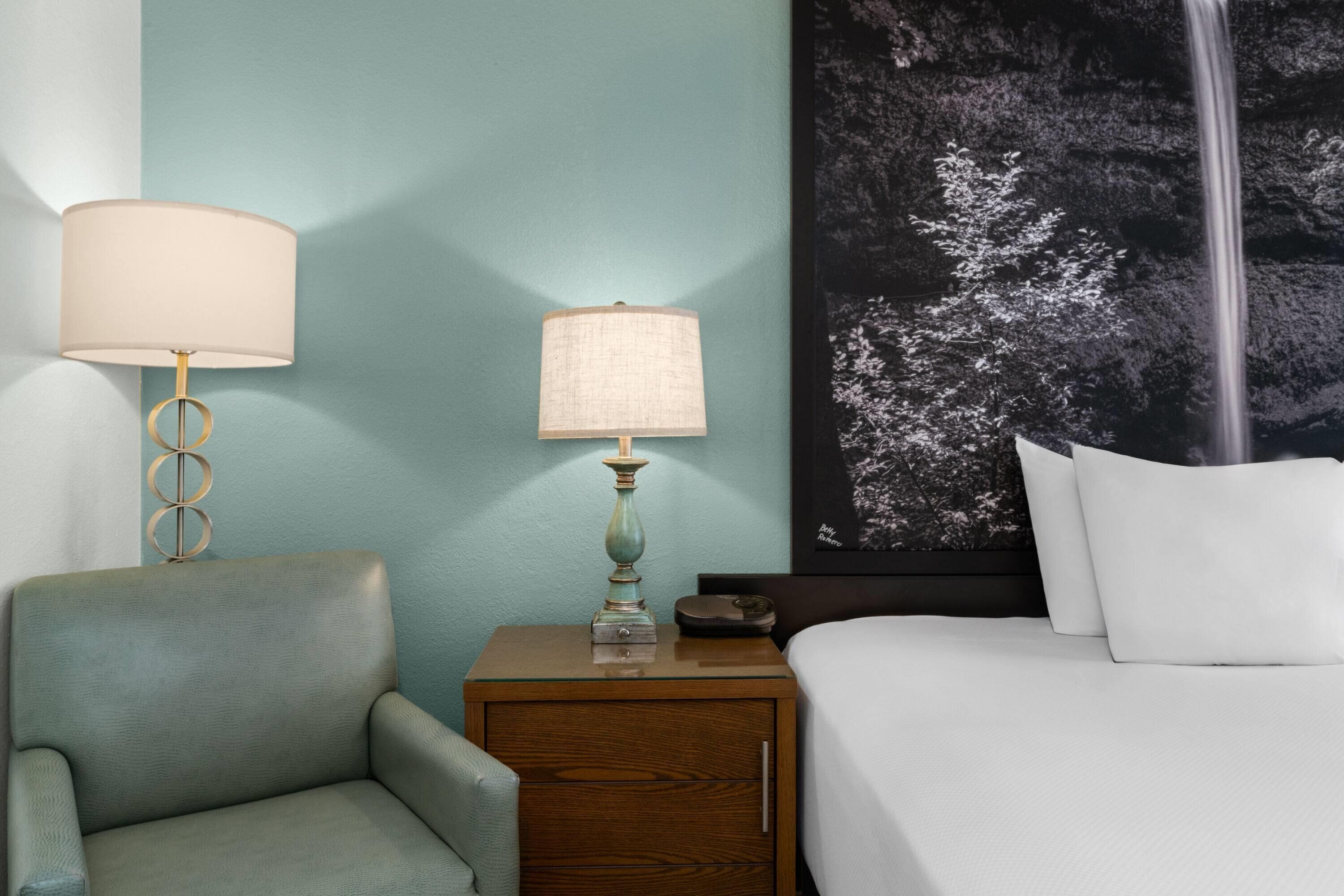 Habitación de hotel con cama, mesita de noche con lámpara, una silla y una obra de arte colgada en la pared.