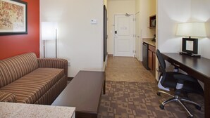 A hotel room with a striped sofa, a small table, a desk with a chair, a lamp, and a door leading to a hallway. - Holiday Inn Lincoln Southwest by IHG (Lincoln)