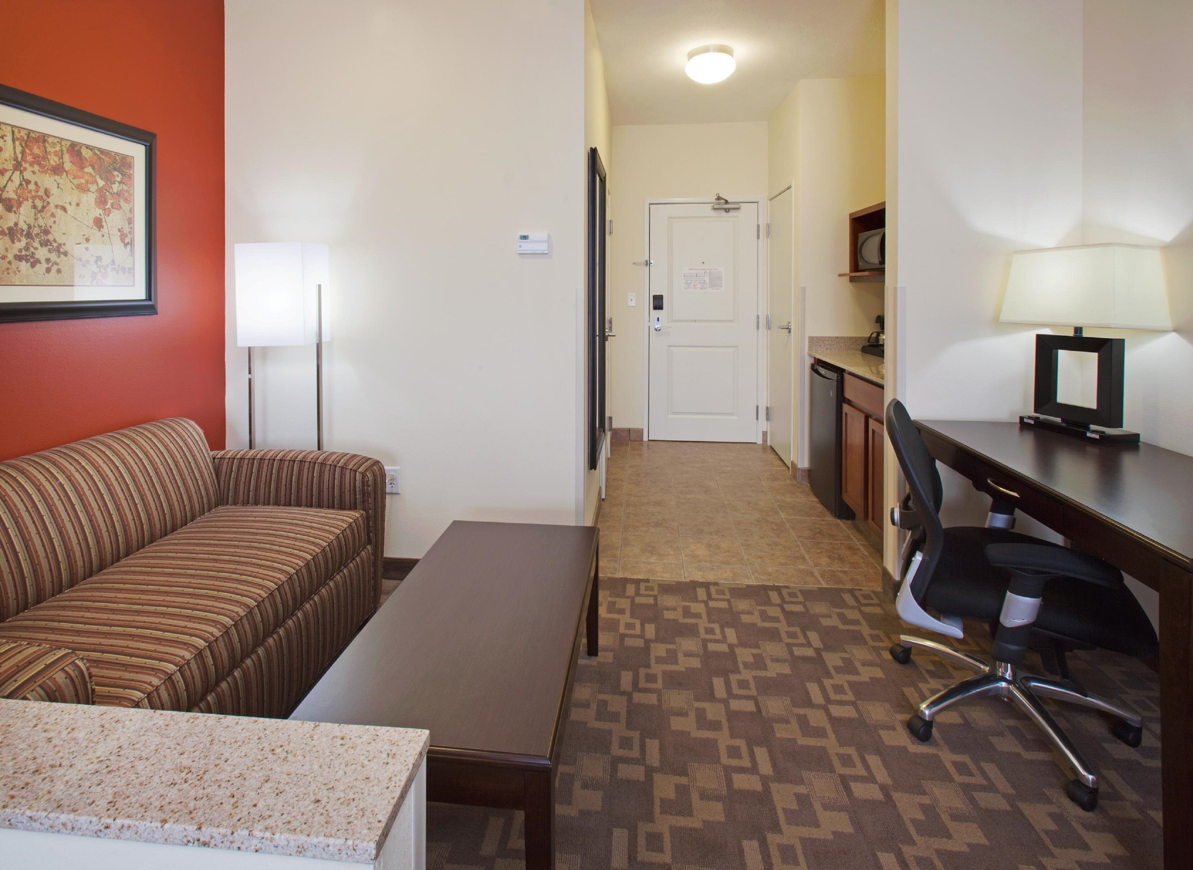A hotel room with a striped sofa, a small table, a desk with a chair, a lamp, and a door leading to a hallway.