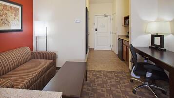 A hotel room with a striped sofa, a small table, a desk with a chair, a lamp, and a door leading to a hallway.