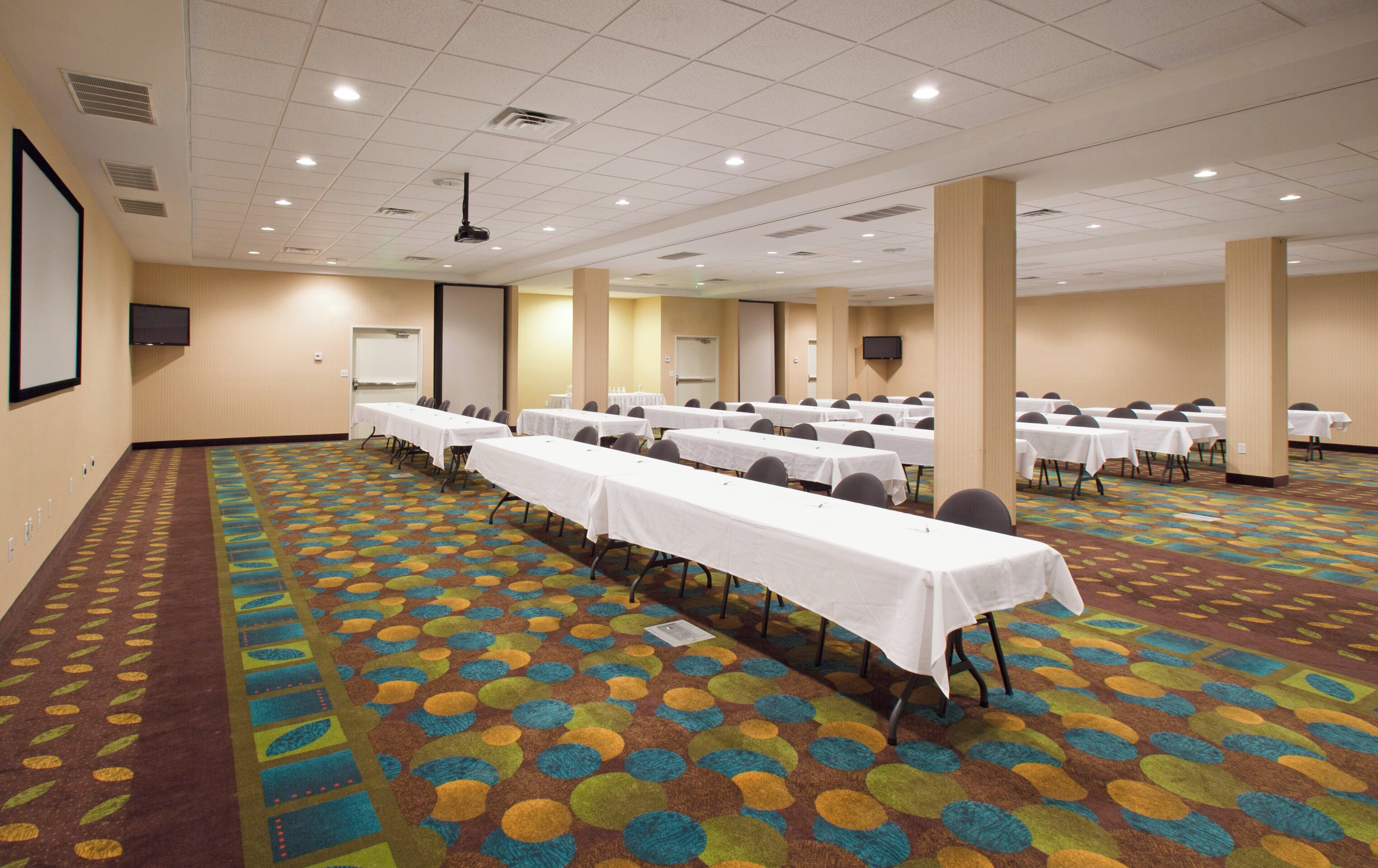 A conference room with multiple tables and chairs, carpeted floor, and a projector screen.