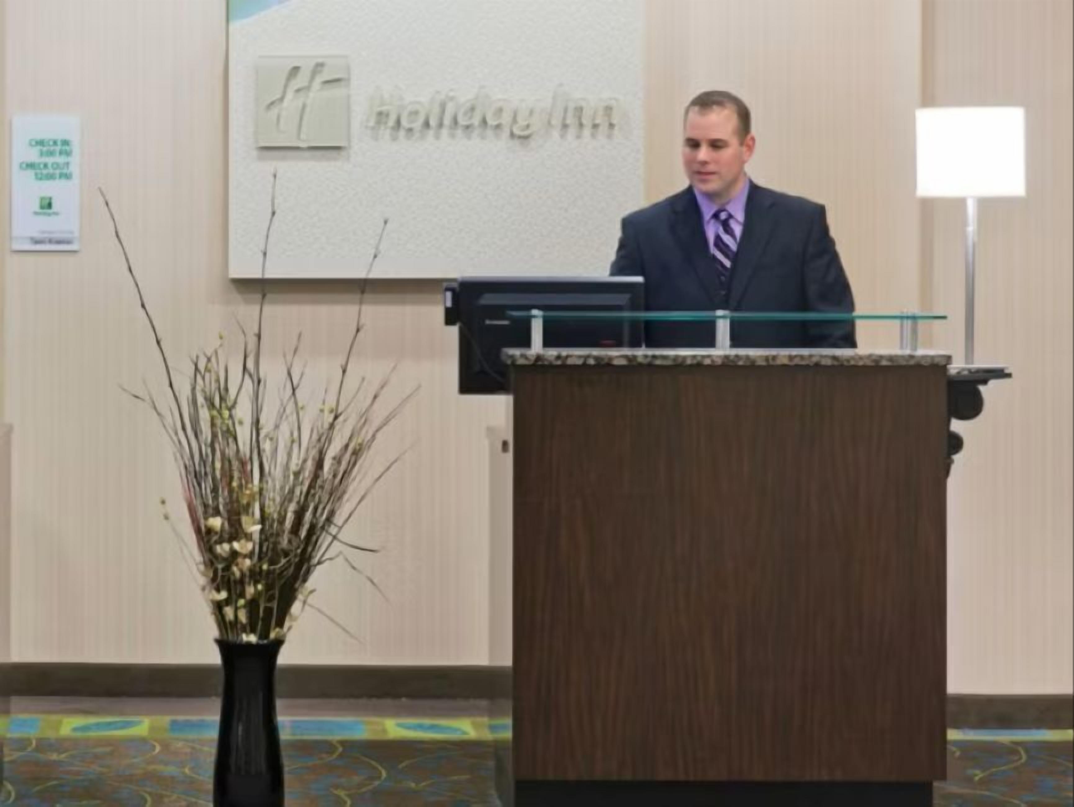 A man at a podium in a room with a 'Holiday Inn' sign on the wall, a check-in/check-out sign, and a vase of branches.