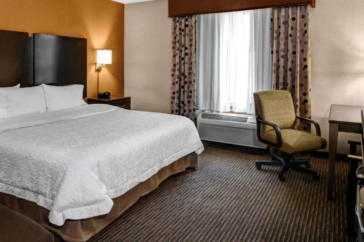 A hotel room with a large bed, a chair, a desk, and a window with curtains.