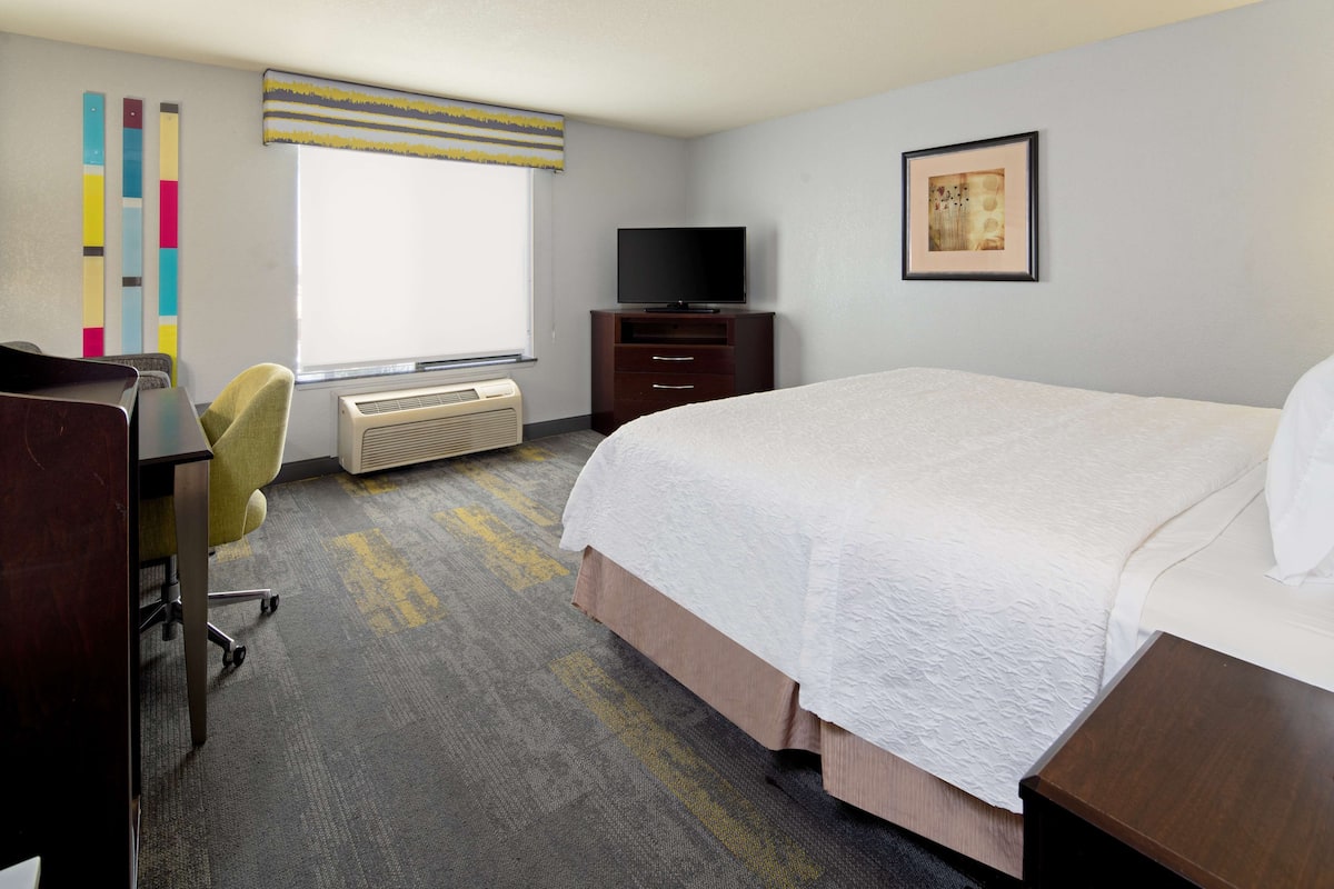 A hotel room with a bed, a desk with a chair, a television, and a window with blinds.