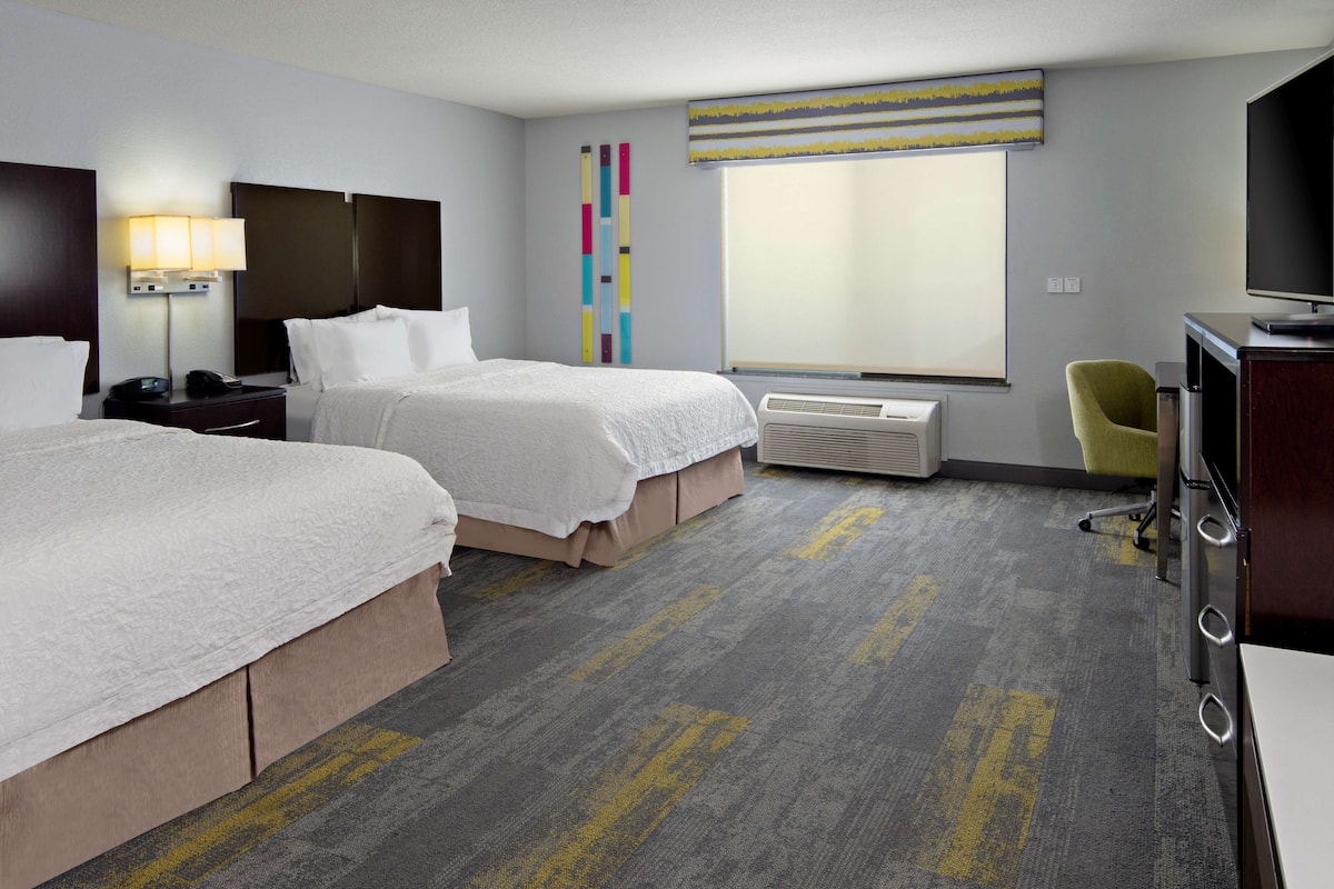 A hotel room with two beds, a desk with a computer, a chair, a large window, and a wall-mounted air conditioner.