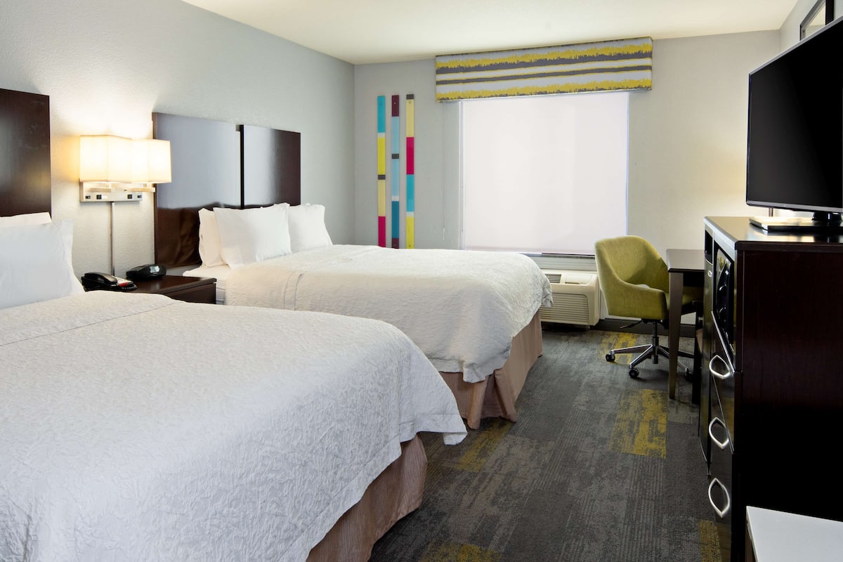 A hotel room with two beds, a desk with a television, a chair, and a window with blinds.