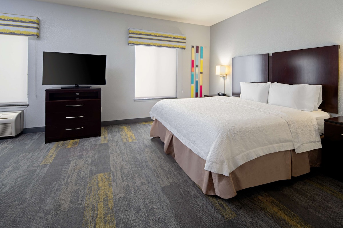 A hotel room with a large bed, a TV, a desk, and a window with blinds.