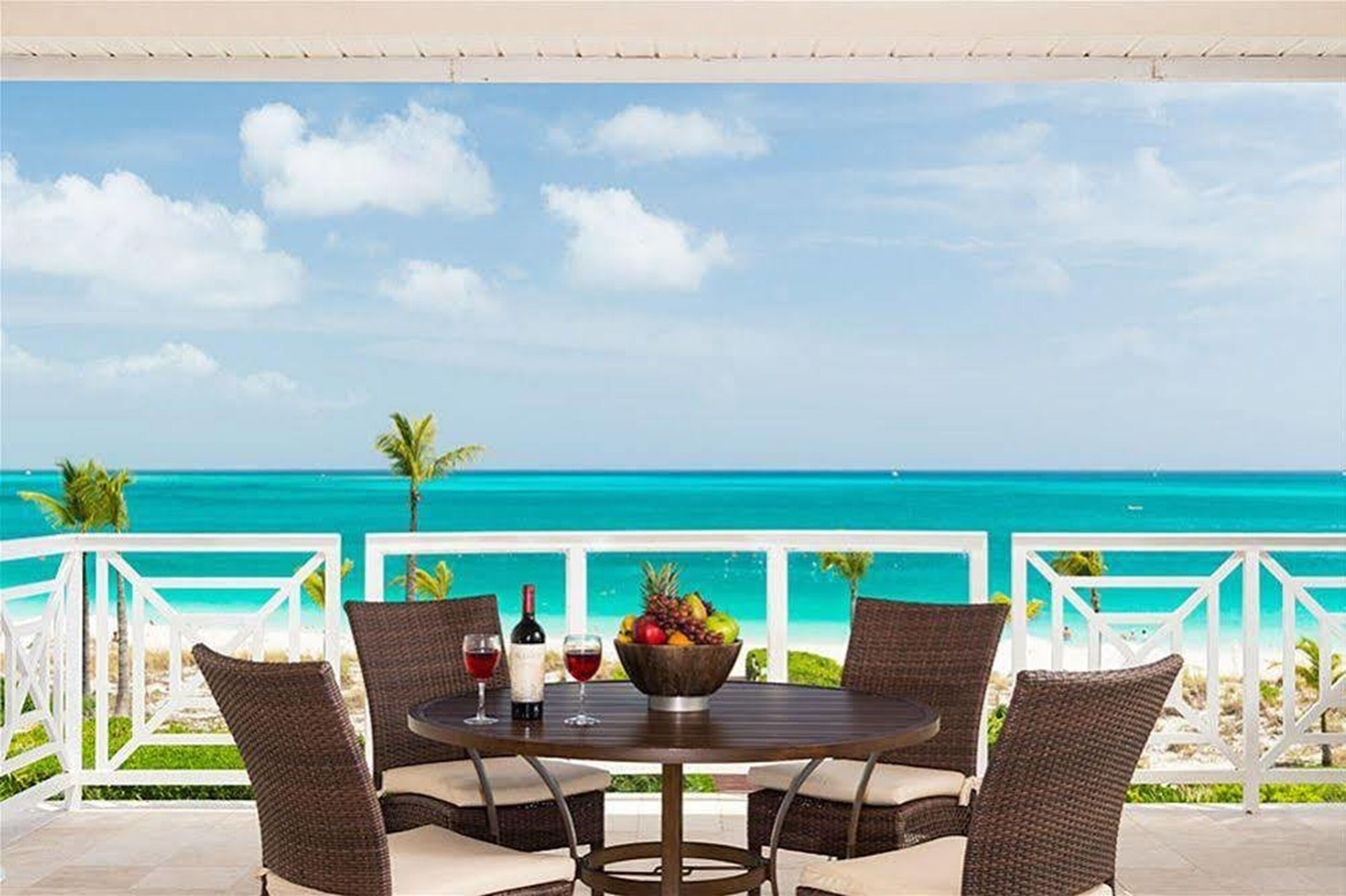 A round table with wicker chairs, a bowl of fruit, and two wine glasses on a balcony with a view of the ocean.