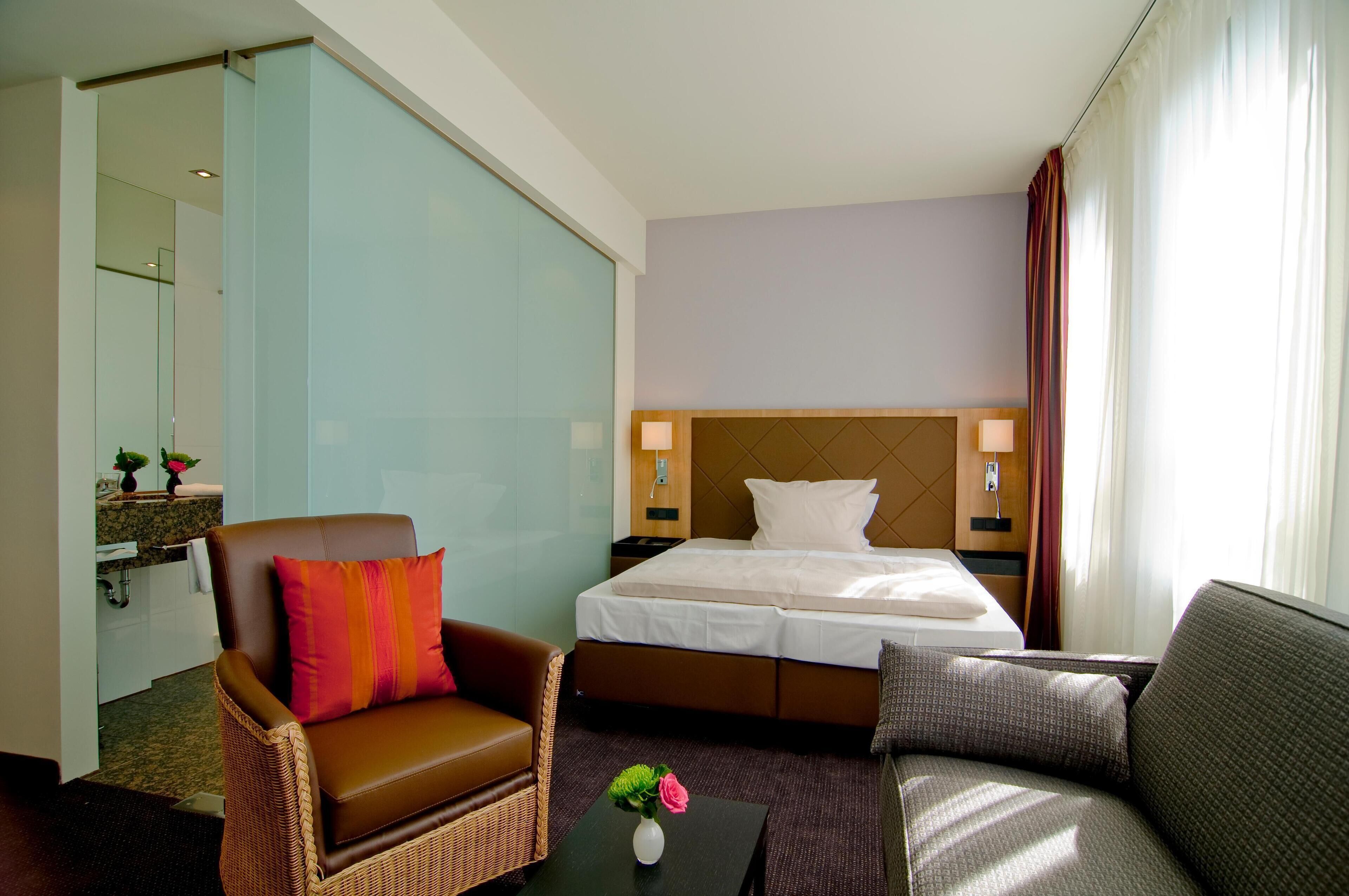 Ein Hotelzimmer mit einem Bett, einem braunen Ledersessel, einem grauen Sofa, einem kleinen Tisch mit Blumenvase und einem großen Spiegel.