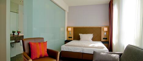 A hotel room with a bed, a brown leather armchair, a grey sofa, a small table with a vase of flowers, and a large mirror.