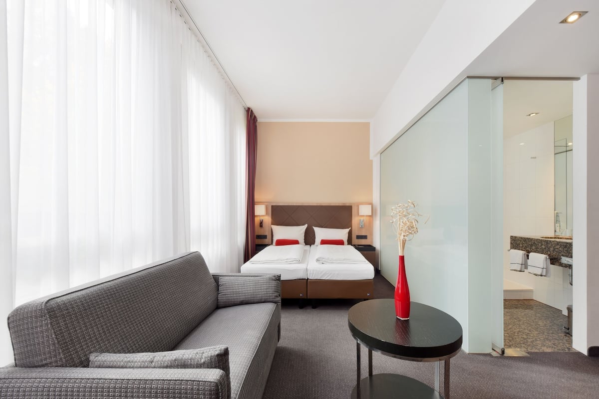 A hotel room with a bed, a sofa, a small table with a vase, and a bathroom visible through an open door.