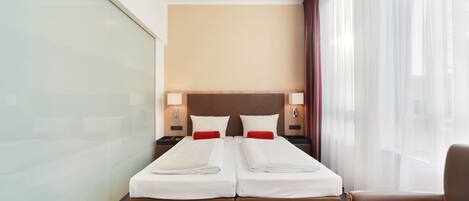 Habitación de hotel con una cama grande, dos mesitas de noche, una silla y vistas a la ciudad a través de cortinas translúcidas.