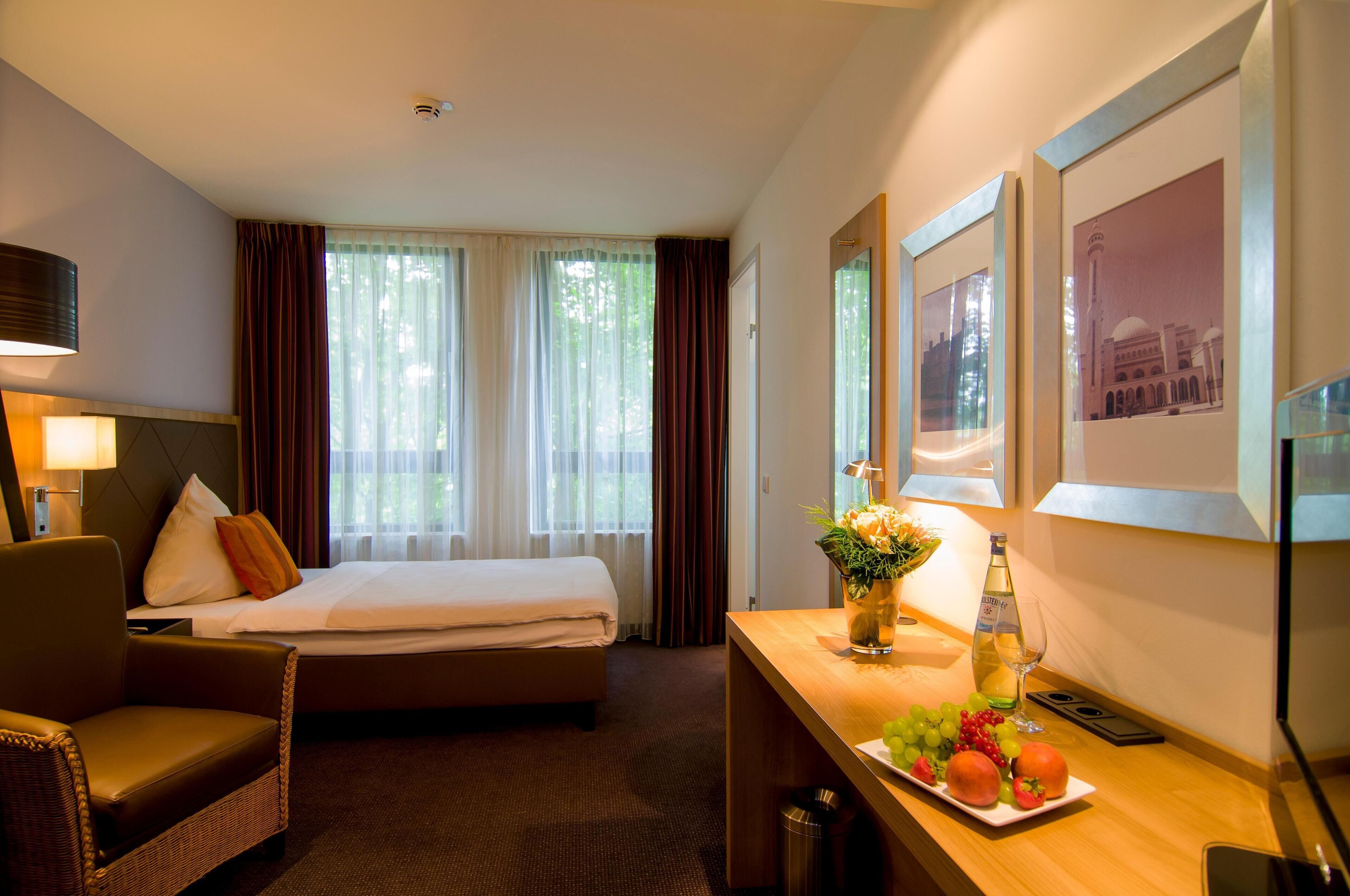 Habitación de hotel con una cama, un escritorio con un florero de flores, un plato de fruta y cuadros en la pared.