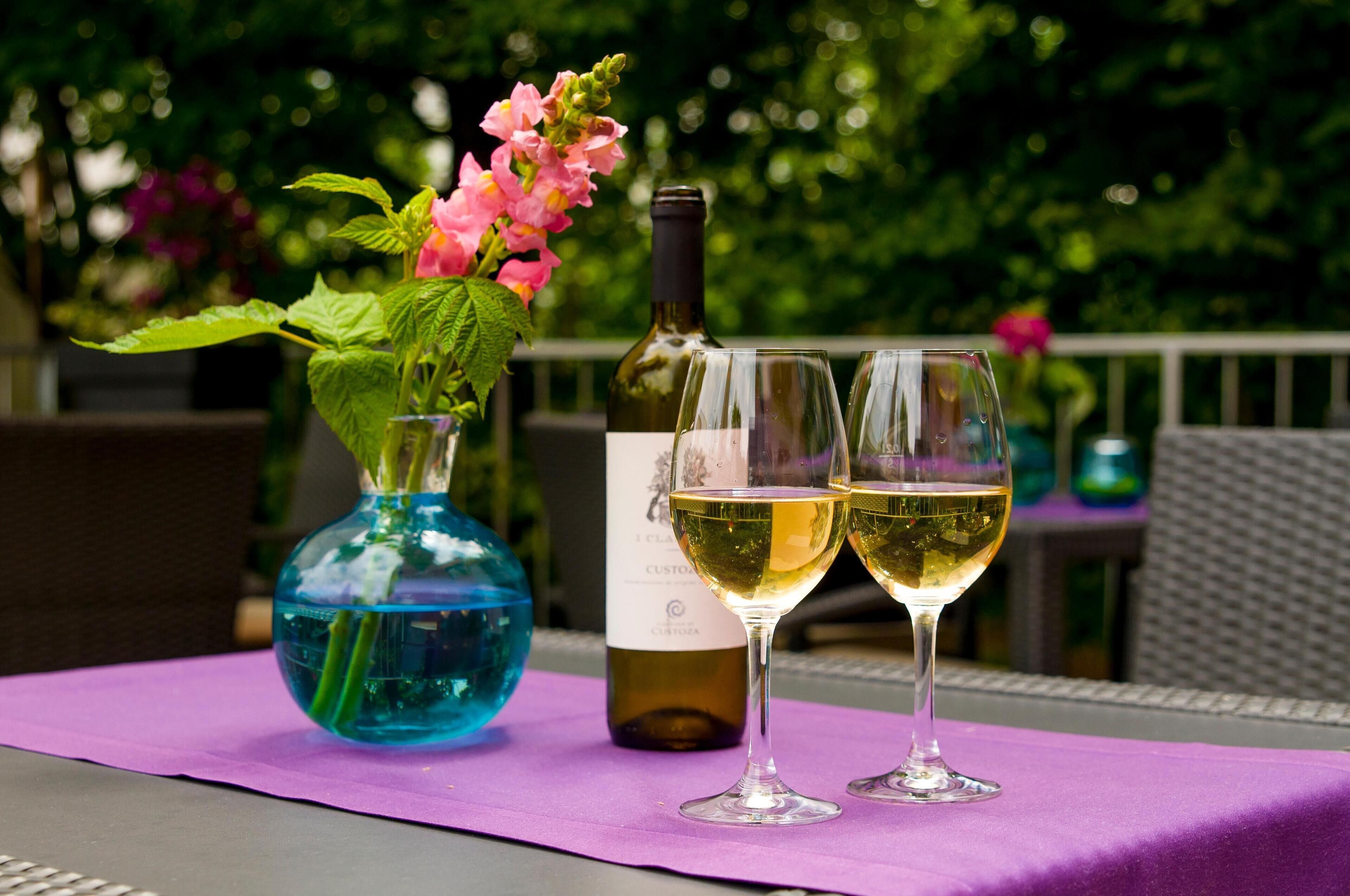 Una mesa con mantel morado, una botella de vino, dos copas de vino y un florero con flores rosas.