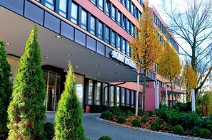 A modern hotel with a glass facade, a covered entrance, and a landscaped garden with coniferous trees. - ACHAT Hotel München Süd (Munich)