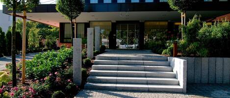 Un edificio moderno con una escalera de piedra que conduce a una entrada de vidrio, rodeado de follaje y un sendero pavimentado.