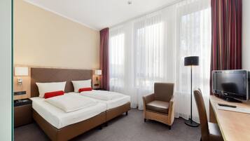 Habitación de hotel con cama doble, escritorio con televisor, una silla y un ventanal con cortinas traslúcidas.