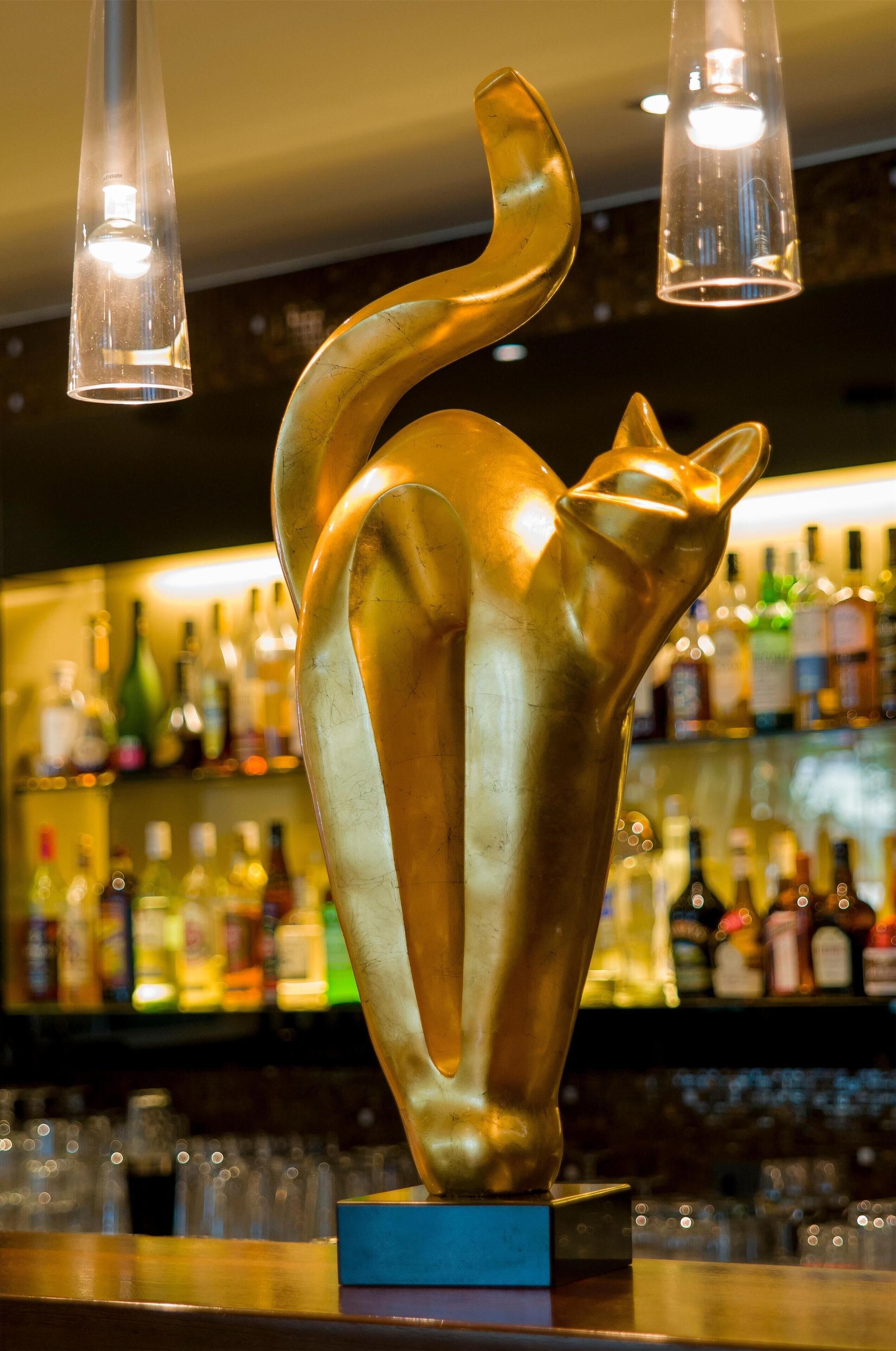 Zona de bar con una escultura dorada de un gato como pieza central, luces colgantes y estantes llenos de botellas de licores.