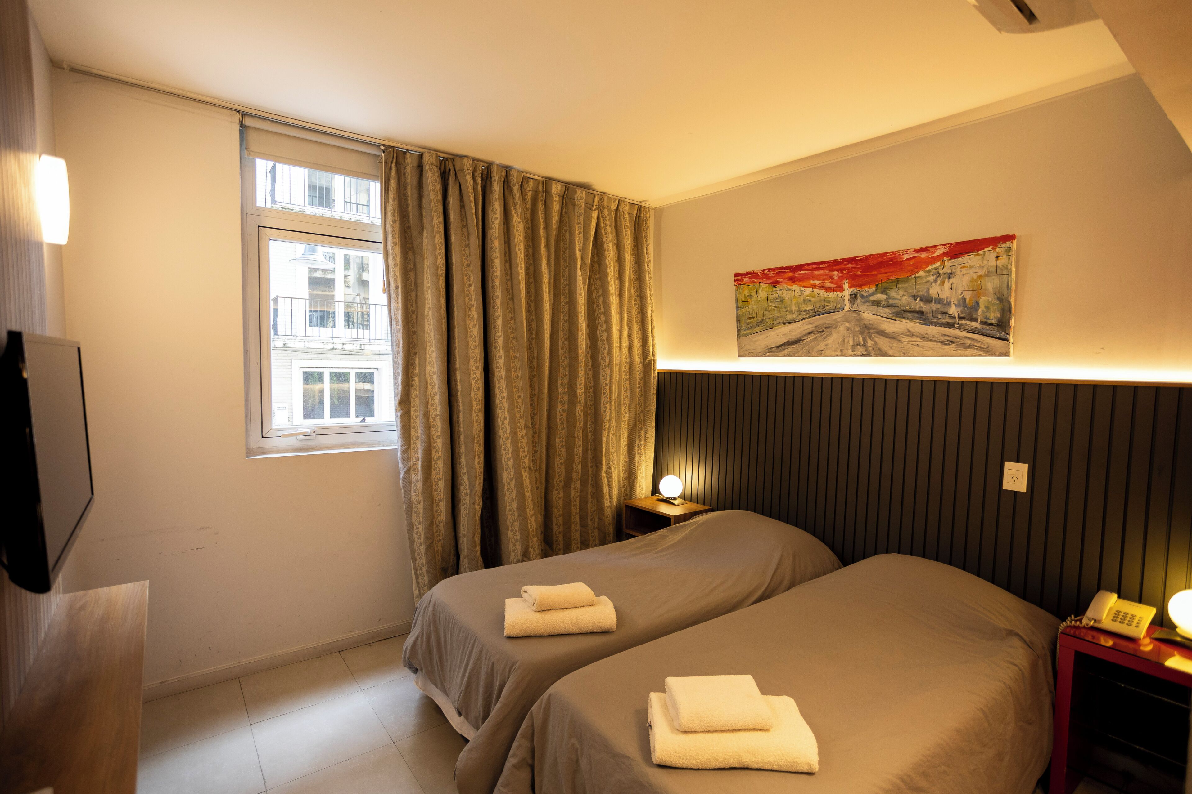 Quarto de hotel com duas camas, um quadro na parede, uma luminária de cabeceira e um telefone na mesa de apoio.