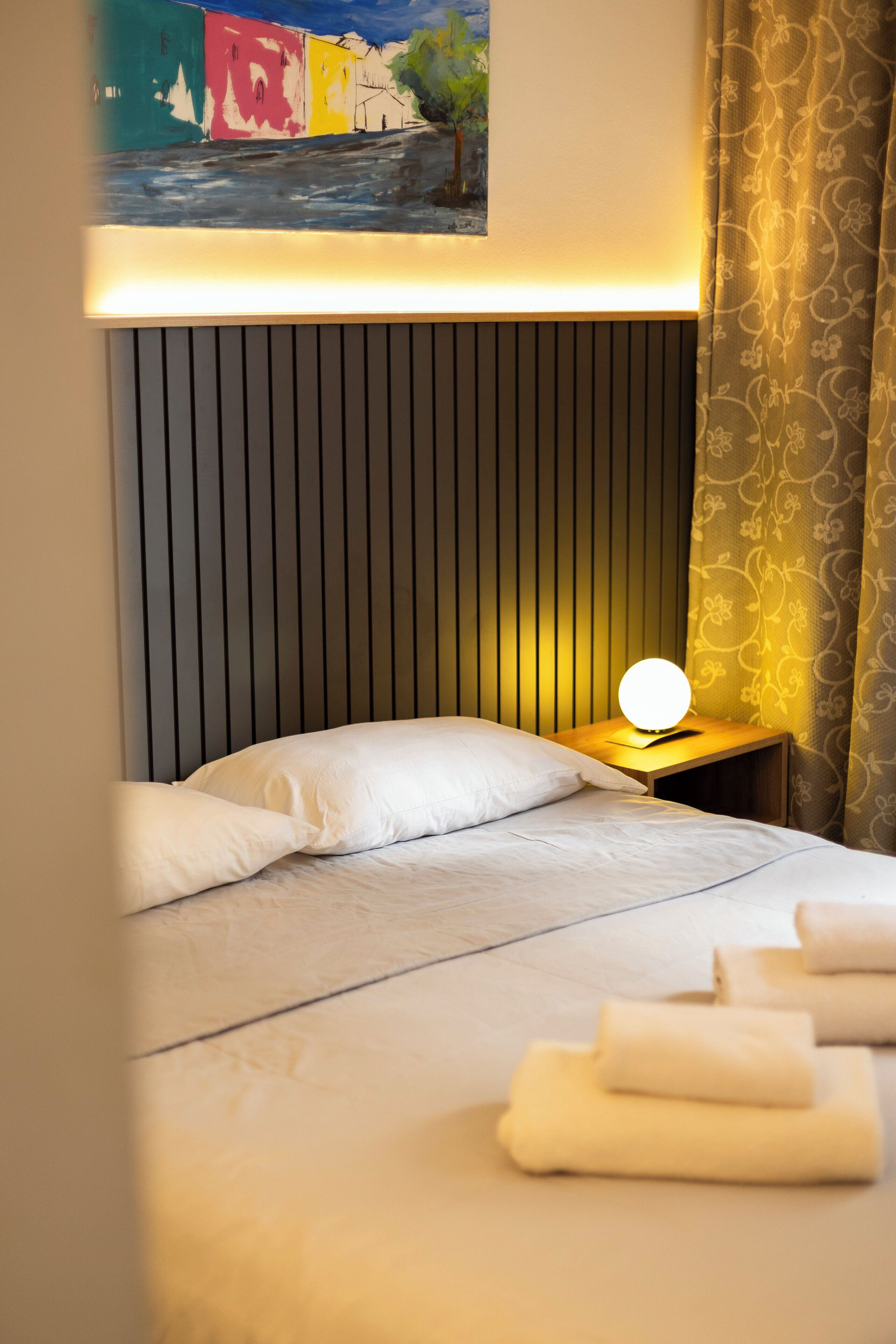 Una habitación de hotel con cama, lámpara de noche, un cuadro en la pared y ventana con cortinas.
