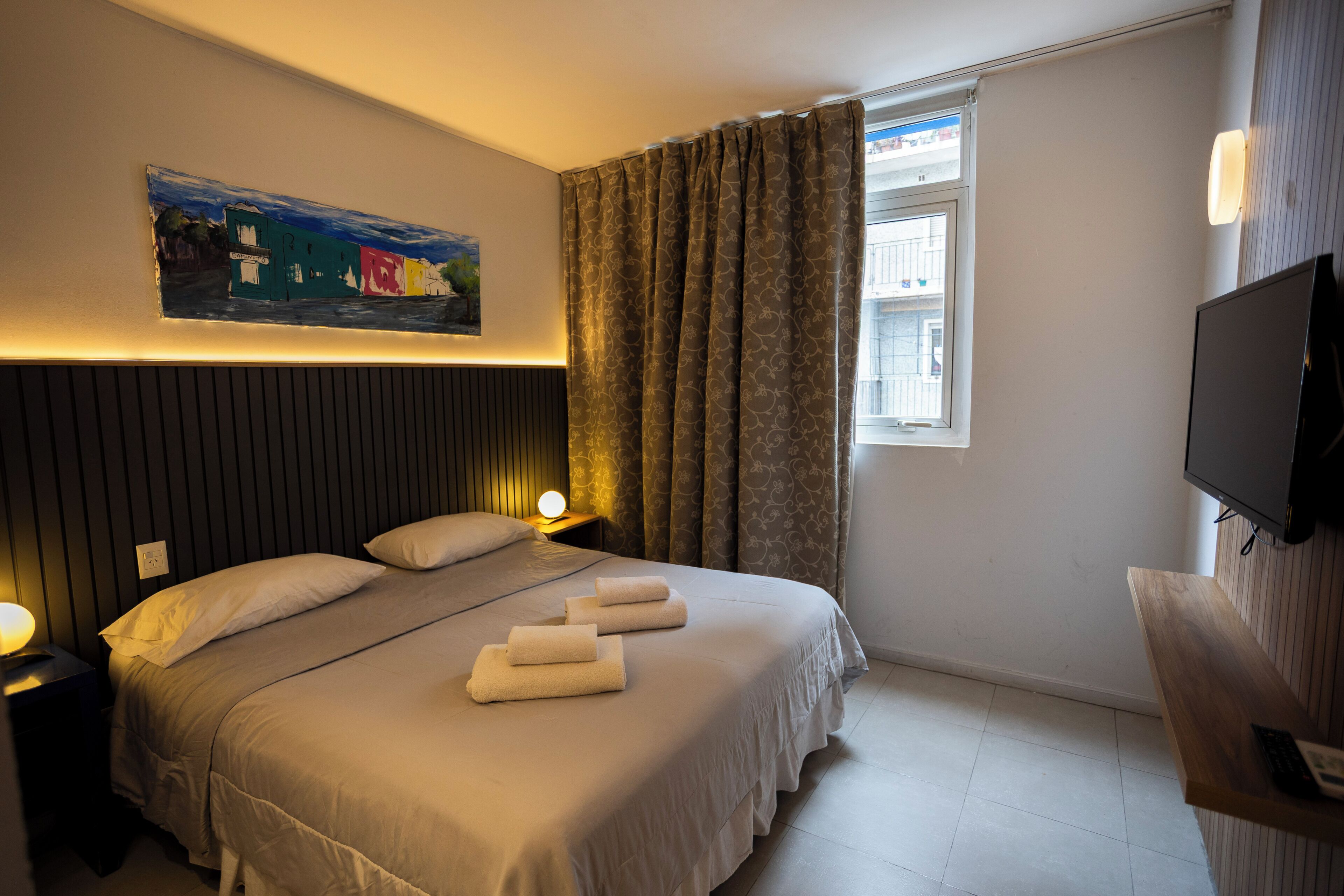 Quarto de hotel com cama, quadro, janela com cortinas, televisão e escrivaninha de madeira.