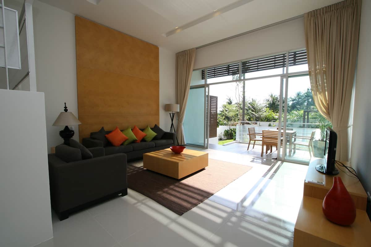 A modern living room with a sofa, coffee table, and a TV. There is a large window with curtains and a view of the outdoors.
