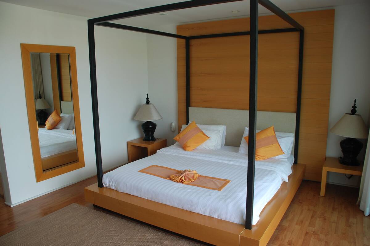 A four-poster bed with white linens and orange accents, a wooden nightstand with a lamp, and a mirror on the wall.