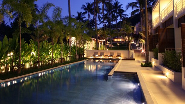 A swimming pool with lounge chairs and a wooden deck, surrounded by palm trees and a building with illuminated windows. - The Park Samui (Koh Samui)