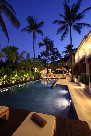 A swimming pool with lounge chairs and a wooden deck, surrounded by palm trees and a building with illuminated windows. - The Park Samui (Koh Samui)