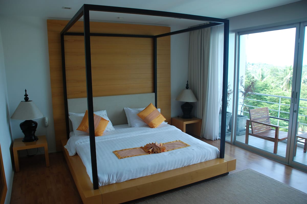 A modern bedroom with a four-poster bed, wooden headboard, and a view of the outdoors through large windows.