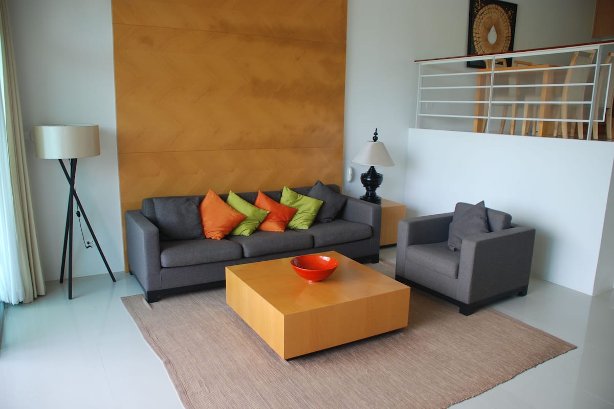 A modern living room with a grey sofa, a smaller grey armchair, a wooden coffee table with a red bowl, and a large orange wall.