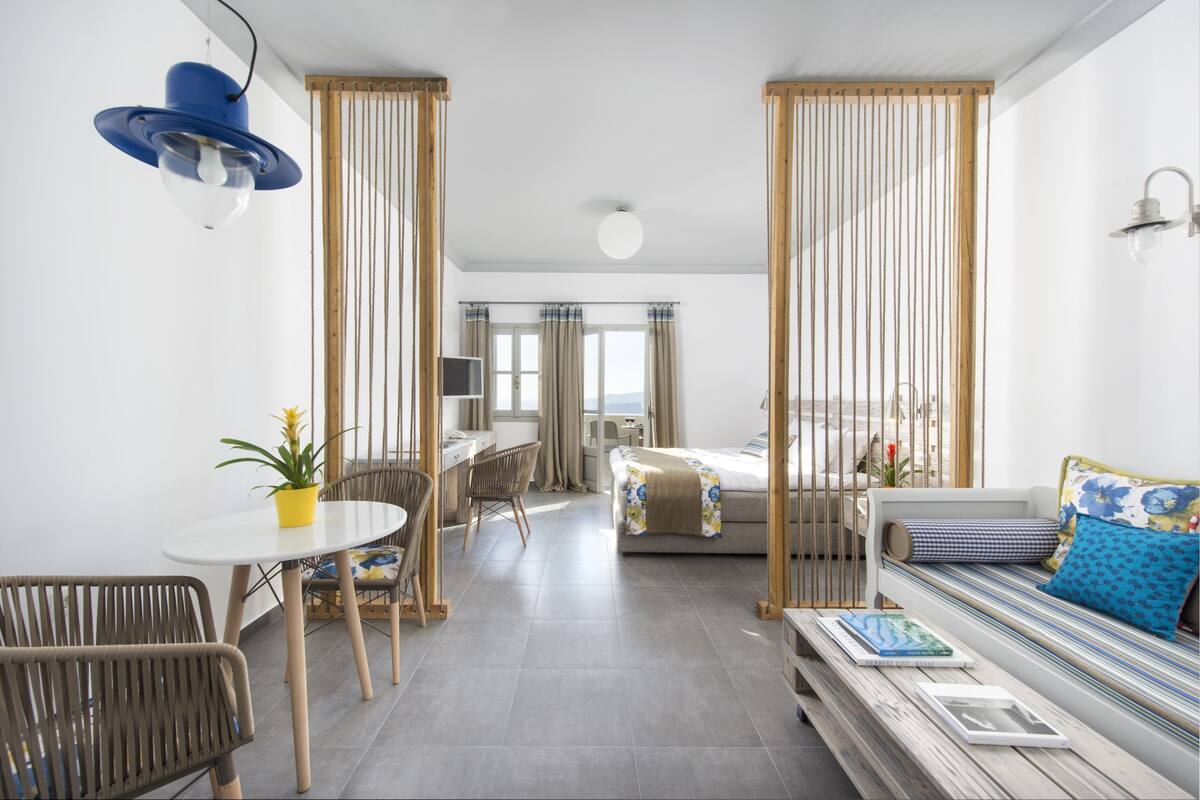A modern hotel room with a bed, a sofa, a dining table with chairs, and a small balcony.