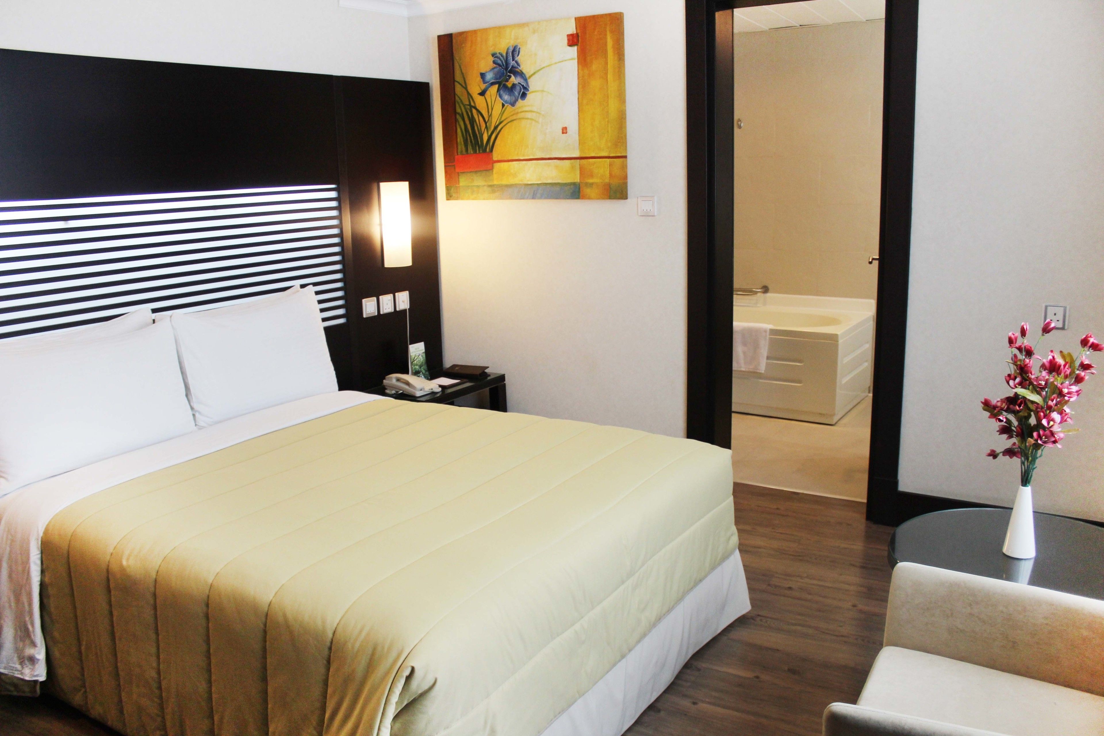 A hotel room with a large bed, bedside table, lamp, and a painting on the wall.