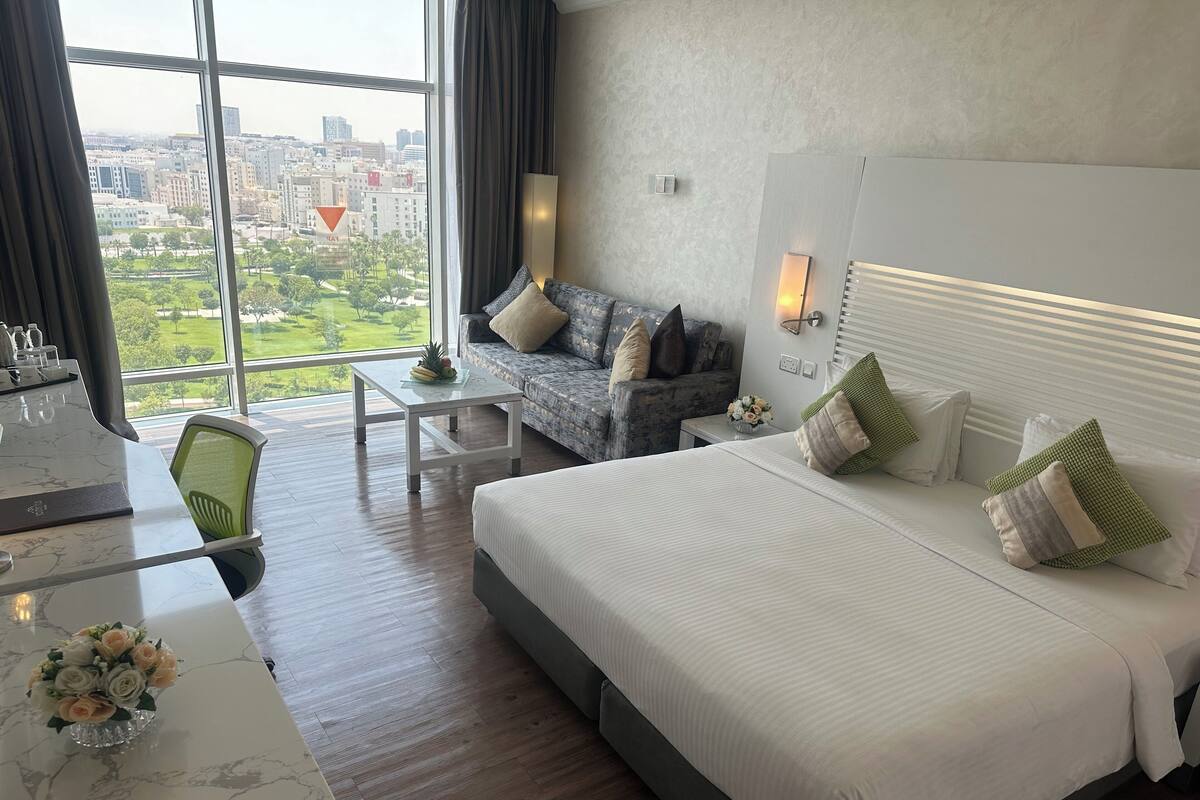 A hotel room with a large bed, a sofa, a small table, and a view of the city through large windows.