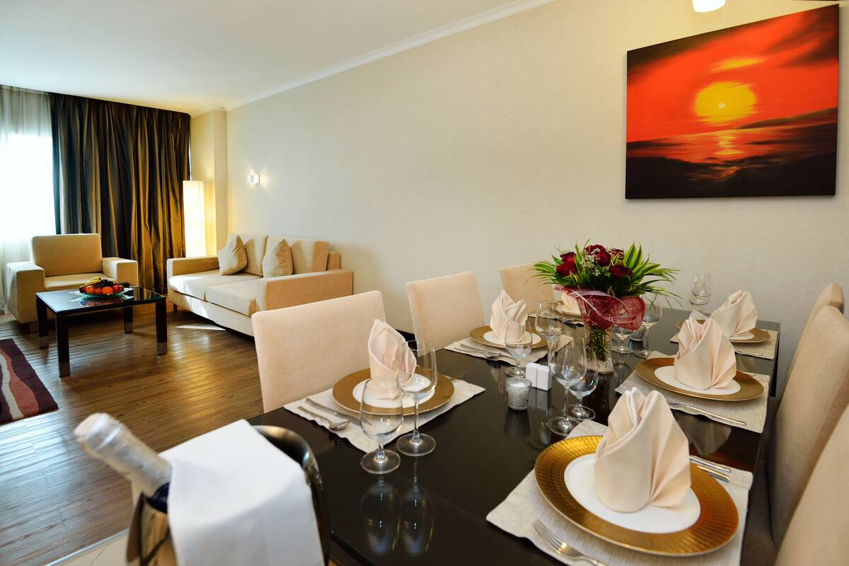 A dining area with a table set for four, including wine glasses, plates, and napkins. There is a painting of a sunset on the wall, and a sofa and armchairs in the background.