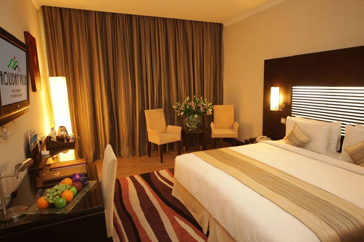A hotel room with a bed, two armchairs, a desk with a lamp, a fruit tray, and a large window with curtains.